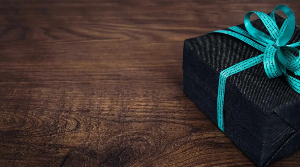 In the picture there is half of a small gift box with a black paper and a light blue ribbon on a wooden table.