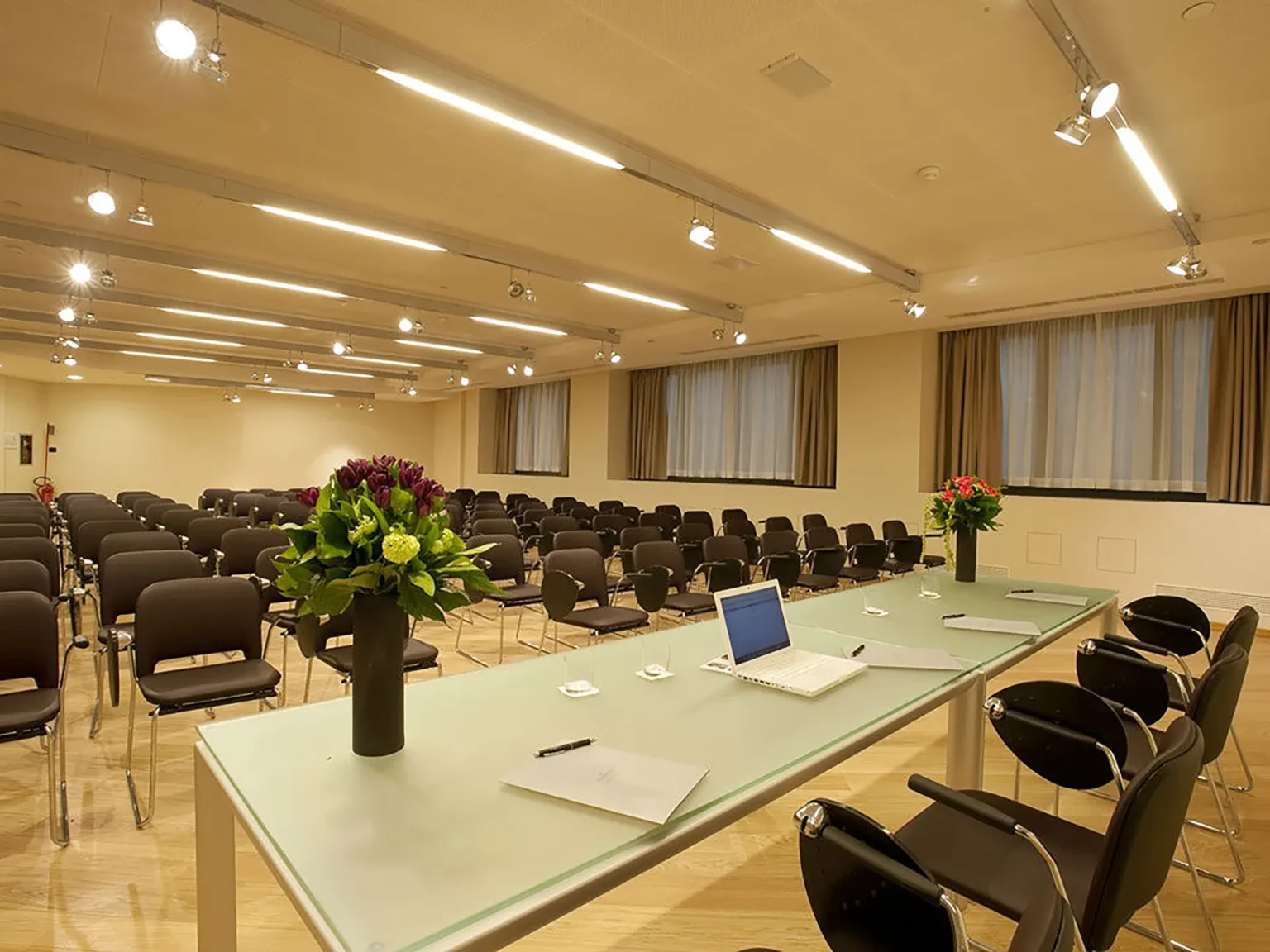 Nell'immagine, la sala meeting del The Hub Hotel: si vede in primo piano un'ampia scrivania con due sedie, sulla scrivania un PC portatile e due mazzi di fiori. Di fronte, alcune file di sedie. Sulla destra, tre finestre con le tende tirate.