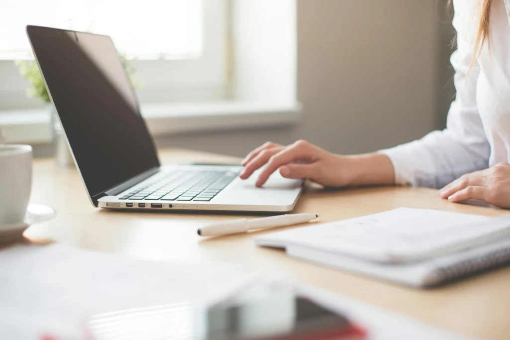 In the picture there is the hand of a girl working on a laptop on a desk. On the right, there is a block notes and a pen and, on the left, a cup.